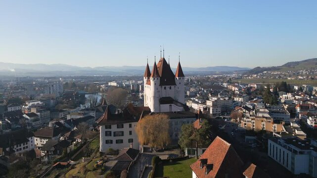 A stunning aerial view of a medieval castle in the historic old town of Thun, Switzerland, surrounded by charming rooftops, a scenic river, and majestic mountains
