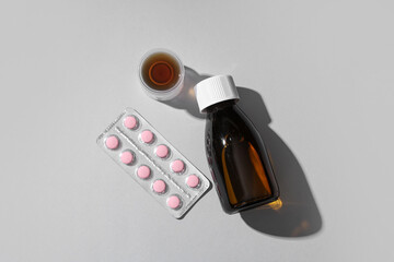 Bottle and measuring cup of cough syrup with pills on grey background