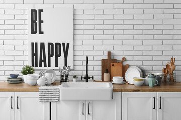 Wooden kitchen counter with ceramic sink, clean dishes and poster hanging on light brick wall