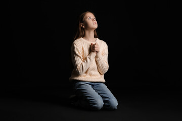 Beautiful teenage girl praying to God on her knees on black background