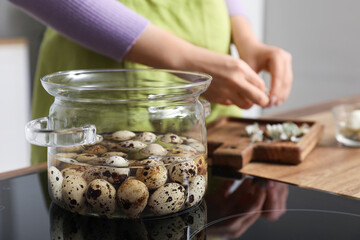 Woman peeling quail eggs in kitchen, focus on cooking pot with boiling eggs