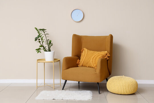 Armchair, pouf. houseplant and clock on white wall in room