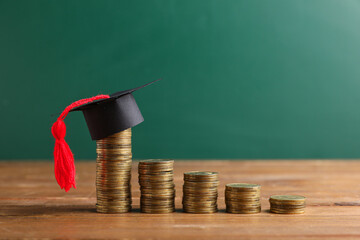 Stacks of coins with graduation hat on table near green blackboard, closeup. Student loan concept