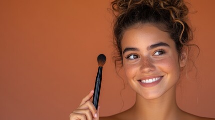 Obraz premium Young woman with curly hair smiles while holding makeup brush against a warm, textured orange background