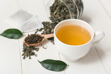 Cup of hot green tea and jar with dried leaves on white wooden background