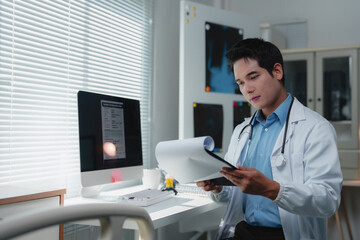 Young male doctor in lab coat and stethoscope reading a medical report on a clipboard in his...
