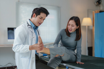 Fototapeta premium Asian male doctor examining injured ankle of young female patient who is experiencing pain while sitting on examination bed in hospital room