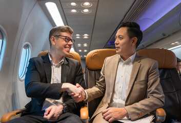 Two businessmen in formal suits shake hands and smile while seated on airplane cabin. Business travel and airline transportation concept