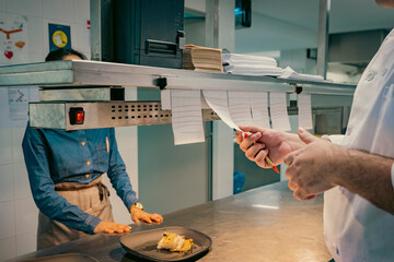 Chef reviewing order tickets in a busy restaurant kitchen. Waiter waits for plated dish. Coordination, teamwork, and efficiency in food service.