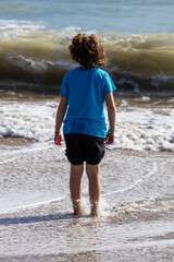 A young boy stands at the ocean's edge, the gentle waves lapping at his bare feet. He's looking out at the waves on a sunny day.