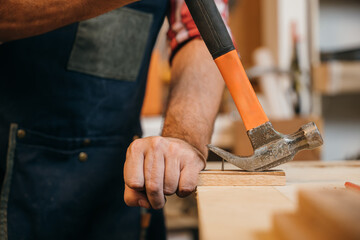A senior carpenter in a workshop filled with tools and supplies uses a hammer to remove rusty nails...