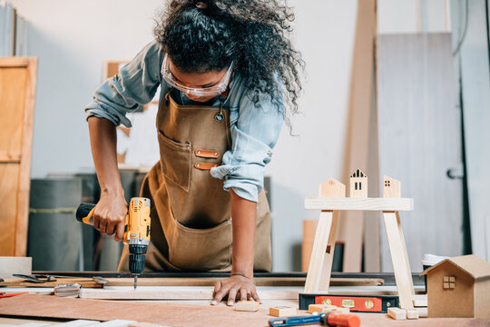 Carpenter woman using a power drill to secure a piece of wood in a workshop, focusing on precision in furniture making. Surrounded by tools and wood pieces, highlighting skill and craftsmanship