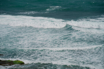 Fototapeta premium Powerful ocean waves crash against the shore, creating white foamy patterns on the deep blue sea. The swirling water and rhythmic waves reflect the beauty and energy of nature.