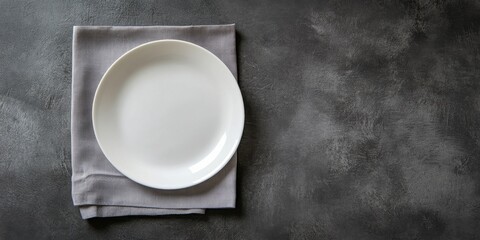 Blank white plate centered on a soft grey napkin against a textured dark backdrop captured from a top view perspective for elegant table setting.