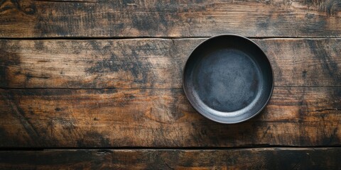 Empty black circular dish centered on a rustic wooden table with a dark grain texture viewed from above creating a minimalist aesthetic.