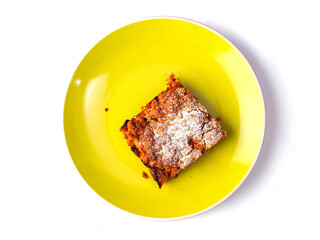 A yellow plate with a piece of cake on it. The cake is covered in powdered sugar. The plate is placed on a white background. Premium slice of a fine bakery product