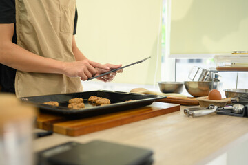 Young man preparing chocolate chip cookies in the kitchen and following recipe on digital tablet