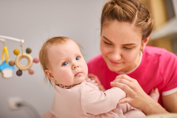 Young mother with brown hair holds baby girl in pink dress indoors. Warm lighting creates intimate, cozy environment. Baby curious expression contrasts with mothers gentle smile