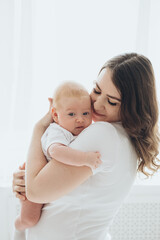 A happy young mother and a newborn baby smile, hug and kiss on a white isolated window background, a loving family at home