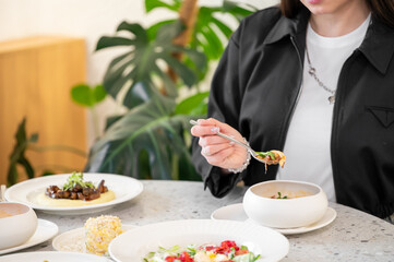 A woman enjoys a meal at a stylish table, holding a fork with a bite of food. Plates featuring diverse dishes are arranged around her, with plants providing a vibrant backdrop
