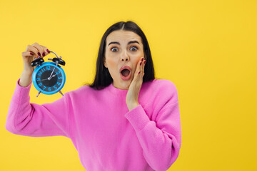 Pretty woman expressing shock while holding alarm clock in studio with yellow background. stressed...