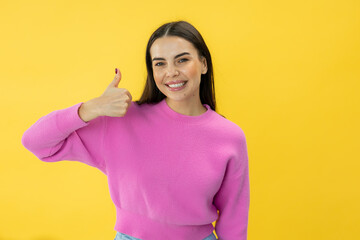 Charming brunette woman with positive smile showing thumb up gesture while posing over yellow studio background. Caucasian young lady in pink sweater giving sincere recommendation and advice.