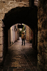 Obraz premium Narrow, atmospheric alley in an old European town Budva, framed by a rustic stone archway. A traveler with backpack stands in the soft morning light, gazing at the historic surroundings.