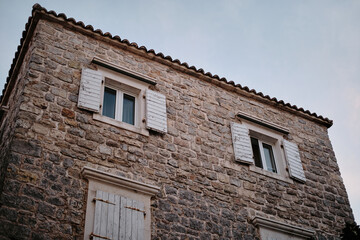 Traditional stone buildings with small windows and wooden shutters in a historic European town. The warm-toned stones contrast beautifully with the clear blue sky. Budva, Montenegro.
