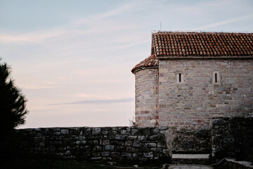 Fototapeta premium Ancient stone building with a red-tiled roof - small chapel or monastery. Pastel sky at sunrise adds a peaceful and timeless atmosphere. Old town Budva, Montenegro.