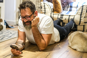 A man of 50 years relaxes at home, lying on the floor and using his smartphone to check social media, surf the web, and make video calls with friends or his companion, while his dog rests beside 