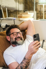Adult man sitting comfortably on the sofa at home, using his smartphone with a relaxed expression...