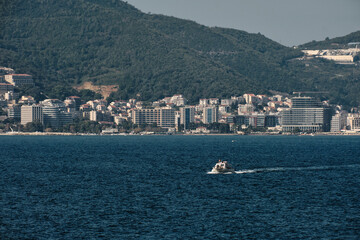 Modern coastal city at the foot of a lush green mountain. Touristic boat glides across the deep blue sea towards the shore, where high-rise hotels and residential buildings line the waterfront.