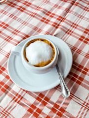 Italian macchiato coffee served in a white cup on a striped red and white tablecloth, evoking midday break rituals, moka brewing traditions, and the cultural charm of after-lunch simplicity
