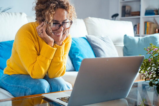 Cozy home interior with a woman sitting comfortably on a sofa, wearing glasses and a yellow sweater, smiling warmly while using her laptop to connect with friends through a video call in her living