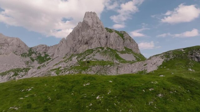 Drone approach to a man standing on a massive rock in the mountains, stopping behind him with a view of the wavy, layered mountains of the Dinaric Alps in Durmitor, Montenegro