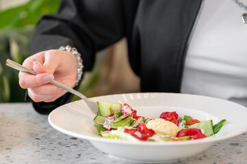 A close-up of a hand holding a fork, eating a fresh salad in a white bowl. The salad contains colorful vegetables and is placed on a speckled grey table