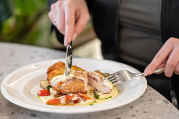 A close-up of a person cutting into a crispy chicken dish on a plate, featuring vibrant vegetables and a creamy sauce, set on a stylish table