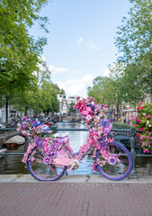 A brightly decorated bicycle covered in pink and purple flowers rests on a canal bridge in Amsterdam. Surrounded by lush greenery and classic canal houses, this eye catching display captures the city