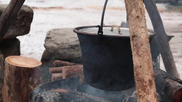 dettaglio di una grande pentola di metallo in primo piano, che contiene della polenta, mentre viene riscaldata su un fuoco, all'aperto, in inverno