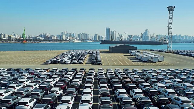 Thousands of brand-new cars for export are neatly parked at Wolmi Port, Incheon, with the city skyline and docked ships in the background under a clear blue sky - aerial dolly