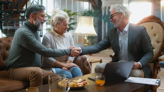 Elderly male insurance agent, sales agent, or real estate agent shakes hands with a retired senior couple after a successful deal at a house meeting. 