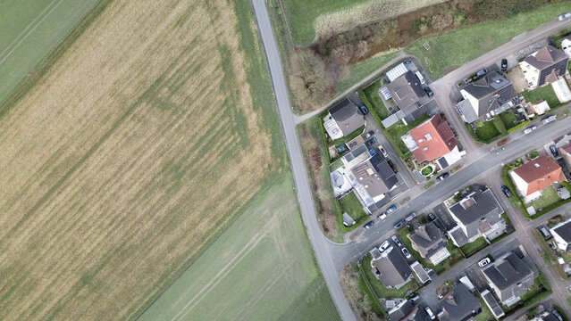 Aerial top-down view of a rural landscape with a paved road separating agricultural fields and a suburban residential area. A contrast between nature and urban development, showing farmland, houses, a