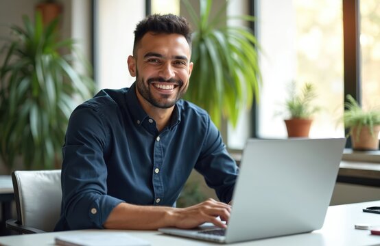 Happy latin businessman works on laptop in office sitting at desk looking at camera. Young entrepreneur smiles using computer tech, cyberspace. Corporate job, lifestyle.