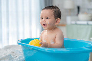 Happy baby enjoying bath time in a blue tub while holding a yellow sponge in a bright, cozy bathroom setting