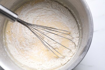 Whisk and bowl of dough on white table, top view