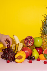 Vertical image of male hand holding a grape next to colorful tropical fruits on a yellow background with pink,pineapple,banana,kiwi,grapes,pear,apple. healthy food