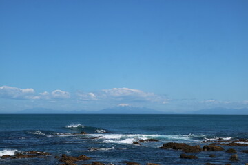 Scenic view of the ocean along the rocky coastline of New Zealand, with gentle waves breaking against the shore. The horizon stretches under a clear blue sky.