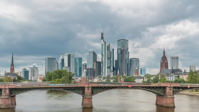 Aerial hyperlapse of Frankfurt skyline with Main River and Ignatz Bubis Bridge. Skyscrapers and church spires reflected in the water beneath dramatic clouds timelapse. Frankfurt, Germany