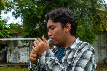 A person in a plaid shirt is lighting a cigarette outdoors, surrounded by greenery and a weathered wall