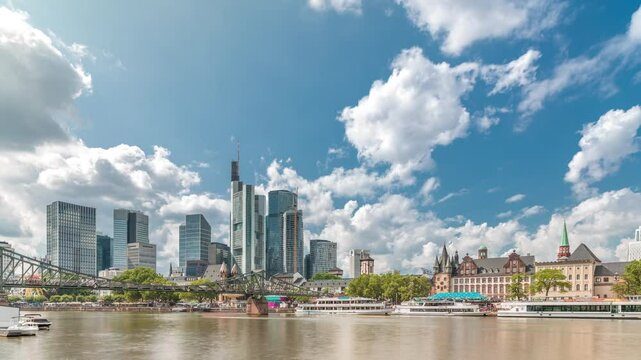 Hyperlapse of the historical Eiserner Steg (Iron Bridge) over the Main River in Frankfurt, Germany. Skyscrapers skyline and historic buildings reflect in the water under a cloud-filled sky timelapse